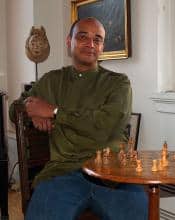 Man in olive shirt sits at a chess table, contemplating a game.