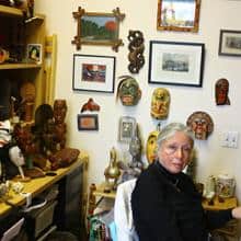 Woman with gray hair sits in a room surrounded by art and artifacts.