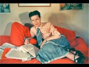 Simone de Beauvoir relaxing on a red couch, surrounded by books.
