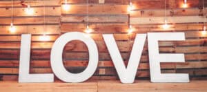 Large white LOVE letters against a rustic wood background with warm string lights.