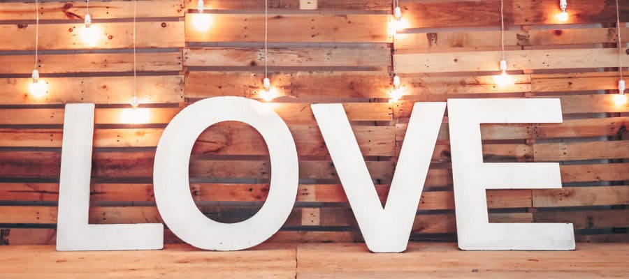 Large white LOVE letters against a rustic wood background with warm string lights.