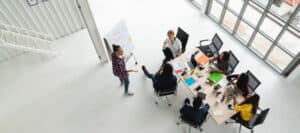 Diverse team brainstorming around a table, one person presenting at a whiteboard.