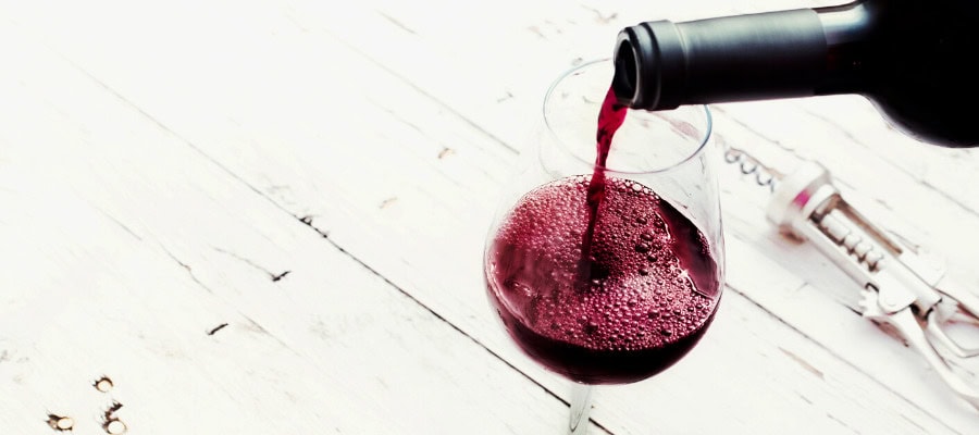 Red wine being poured into a glass on a white wooden table. A corkscrew is nearby.
