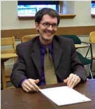 Smiling man in suit sitting at a desk, possibly discussing Kierkegaard.