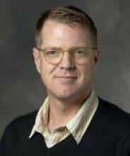 Headshot of a middle-aged man with glasses, wearing a dark v-neck sweater over a collared shirt.