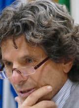 Close-up portrait of a man with curly gray hair and glasses, thoughtfully touching his chin.