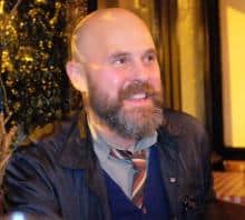 A man with a beard, wearing a jacket and patterned tie, smiles thoughtfully.