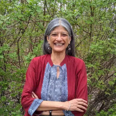 Smiling woman with gray hair wearing a red shirt, standing in front of a leafy green bush.