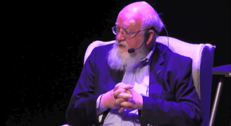 Dan Dennett sits in a chair with hands clasped, wearing a suit and microphone.