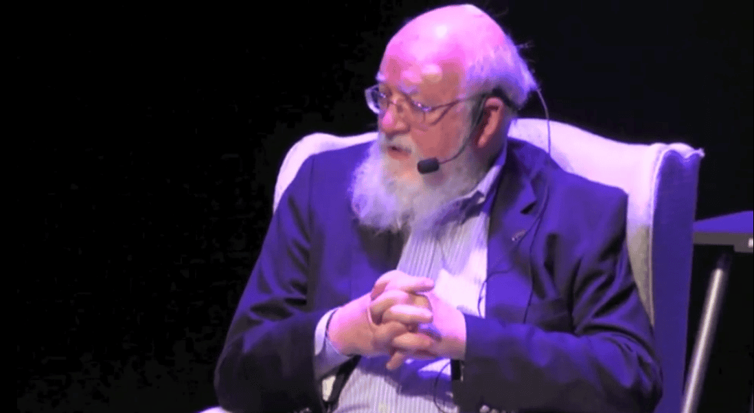 Dan Dennett sits in a chair with hands clasped, wearing a suit and microphone.