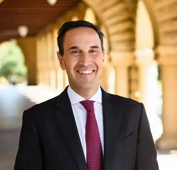 Man in suit smiling in front of Stanford University architecture