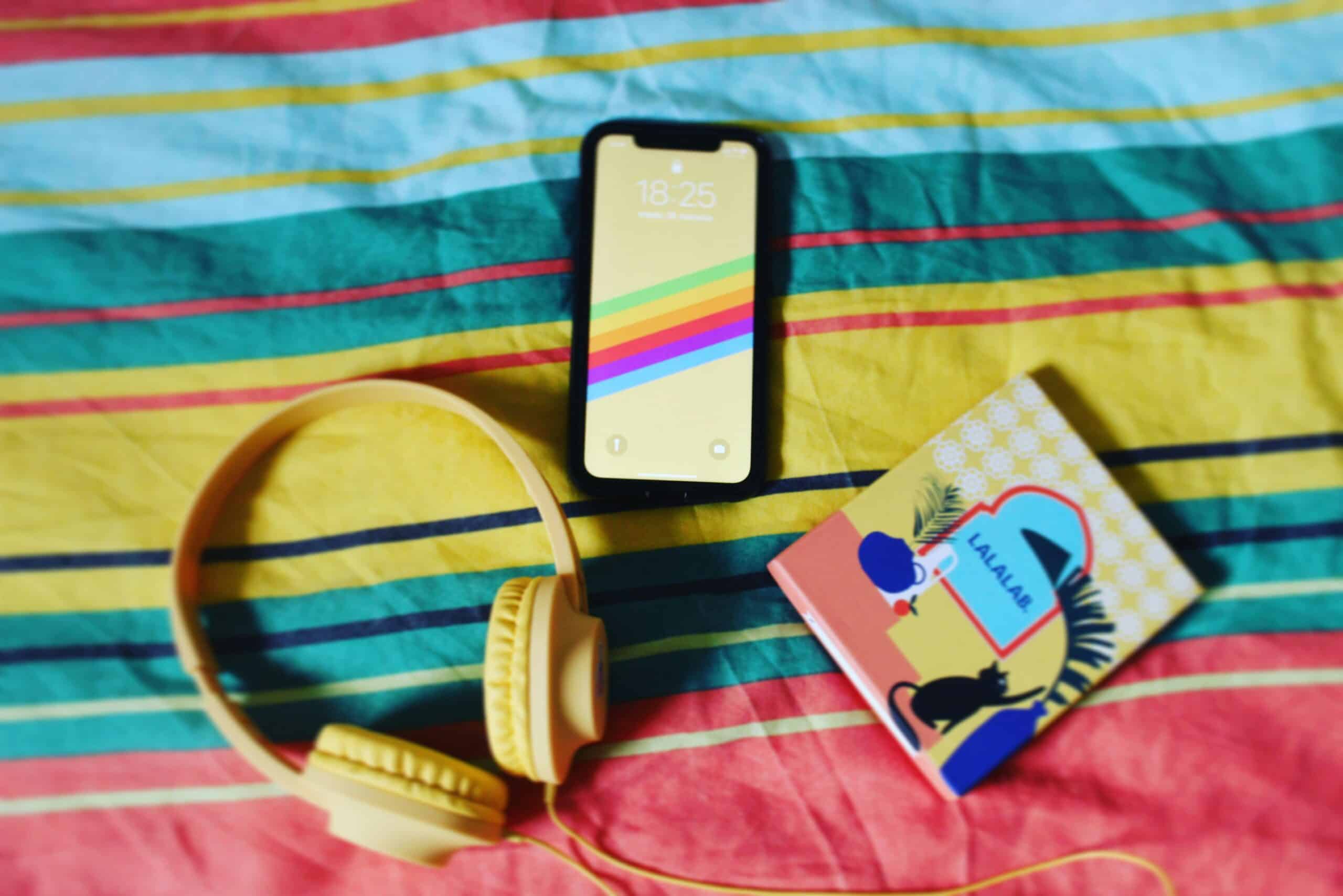 Yellow headphones, phone with rainbow wallpaper, and a notebook on a striped background