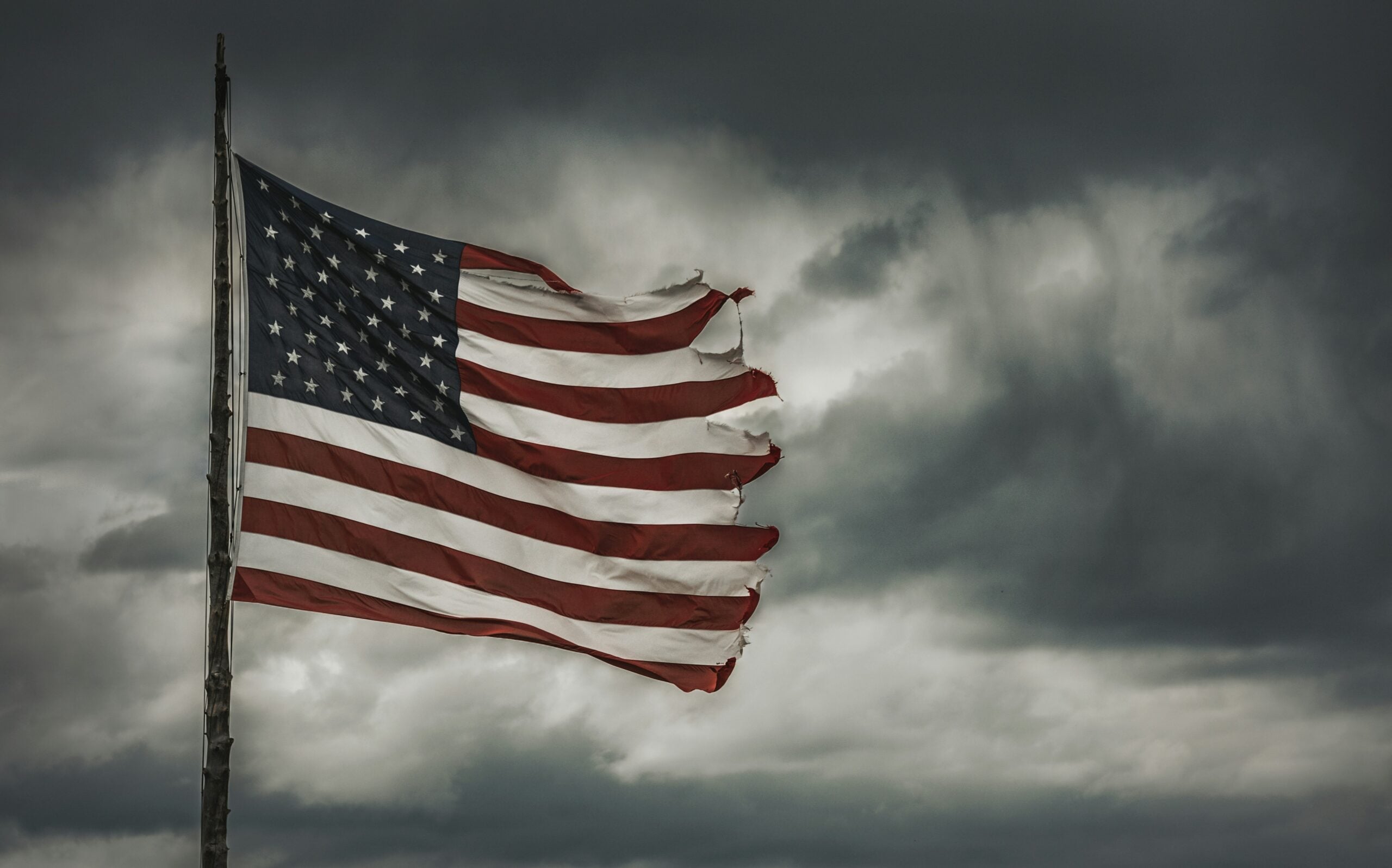 Tattered American flag waves against a stormy, dark sky.