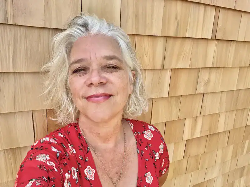 Woman with gray hair and a red floral dress smiles against a wooden wall.