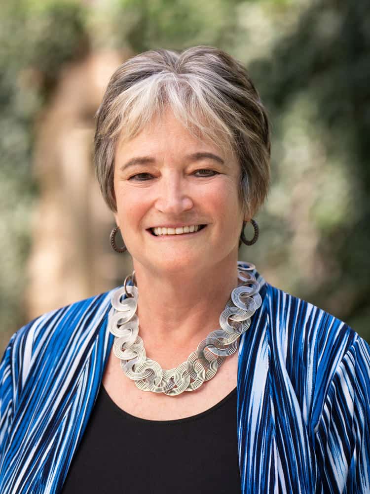 Portrait of a woman with short, graying hair, wearing a blue and white striped jacket and a statement necklace.